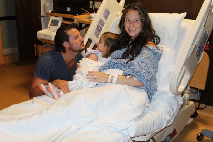 Dad kissing new baby girl and daughter and mom in hospital bed