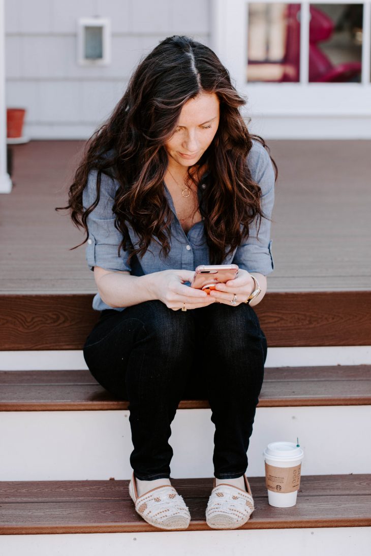 Woman in Blue Shirt on Cell Phone Sitting on Stairs with a Coffee Outside