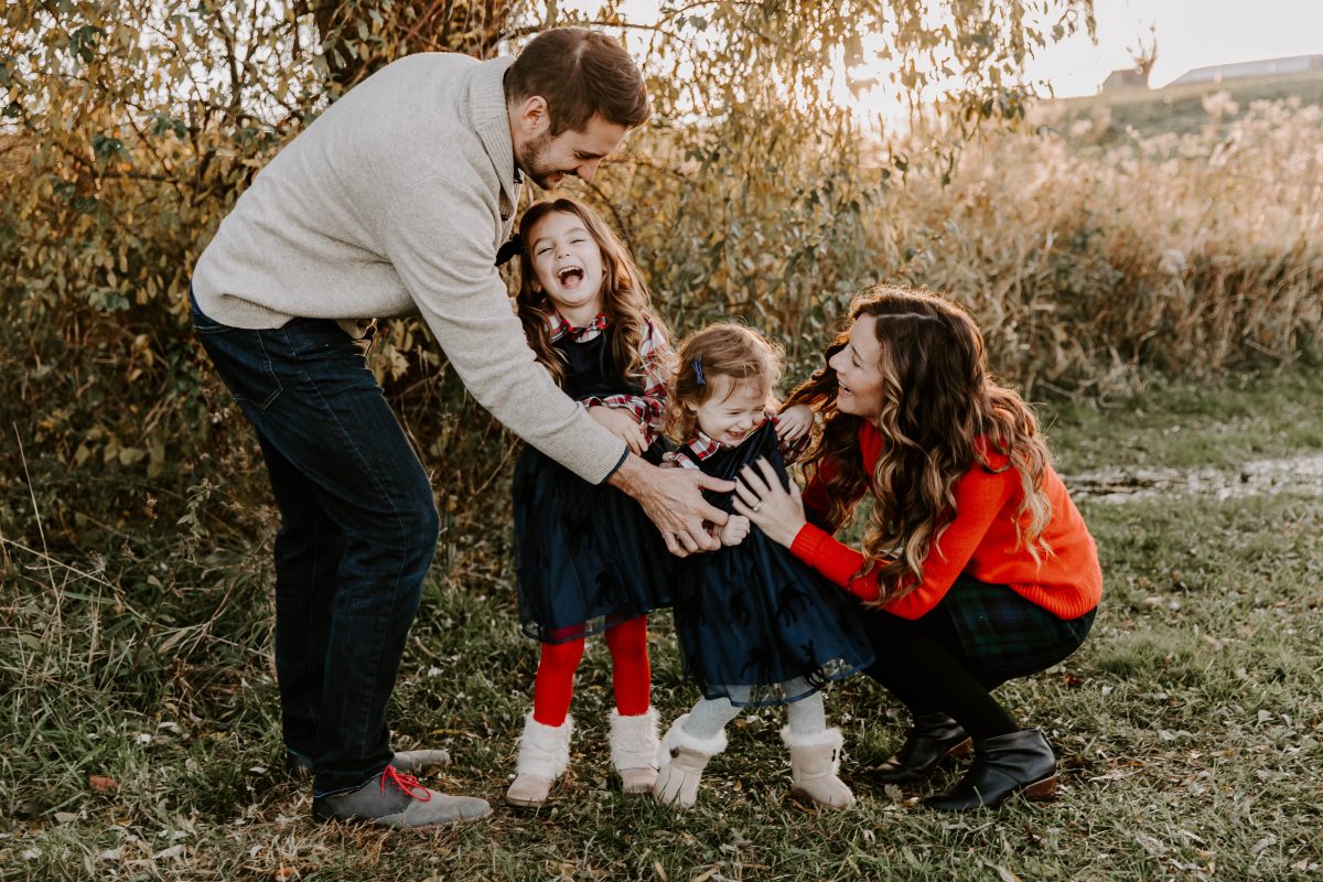 Man in Cream Sweater looking at youngest daughter wearing plaid shirt and navy dress. Child is in Mother's arms and another daughter stands between the parents looking up at them