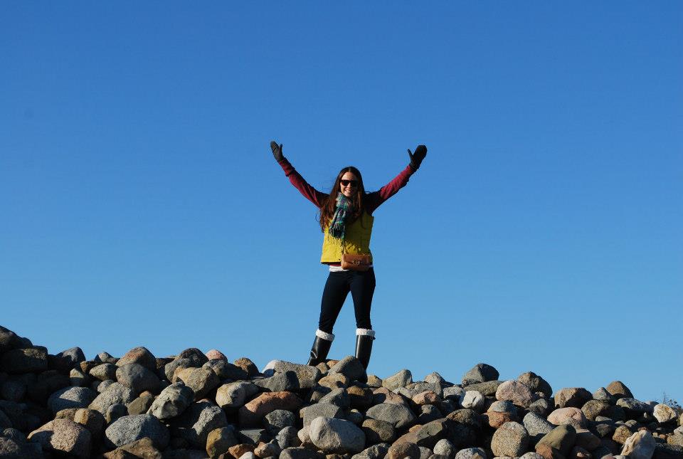 Woman wearing vest, scarf, gloves and boots with arms raised and smiling as she is standing on top of rock pile in Norway