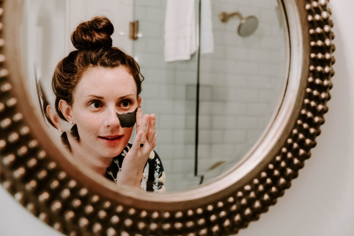 Woman wearing lemon print pajama top applying charcoal mask to her face in front of a mirror