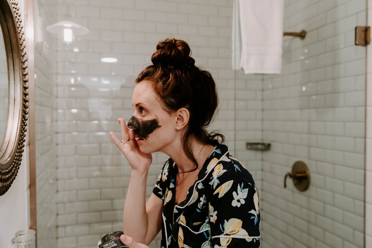 Woman wearing lemon print pajama top applying charcoal mask to her face in front of a mirror