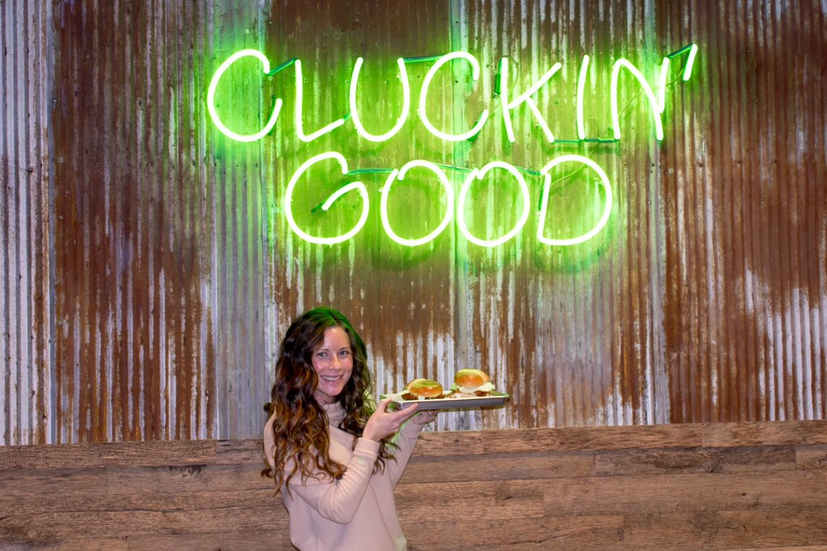 A girl in a tan top holding a tray of chicken sandwiches under a neon sign that says "Cluckin' Good"