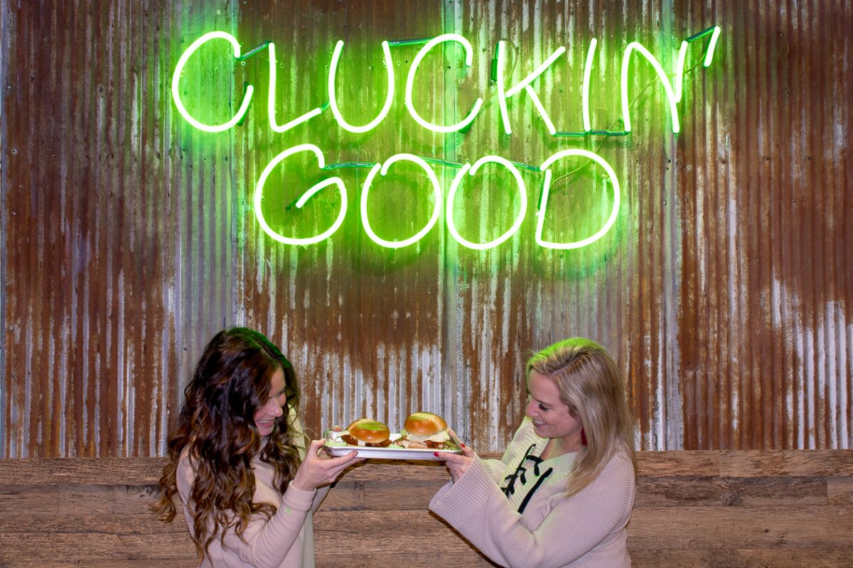 Two girls in tan shirts holding a tray of chicken sandwiches under a neon sign that says "Cluckin' Good"
