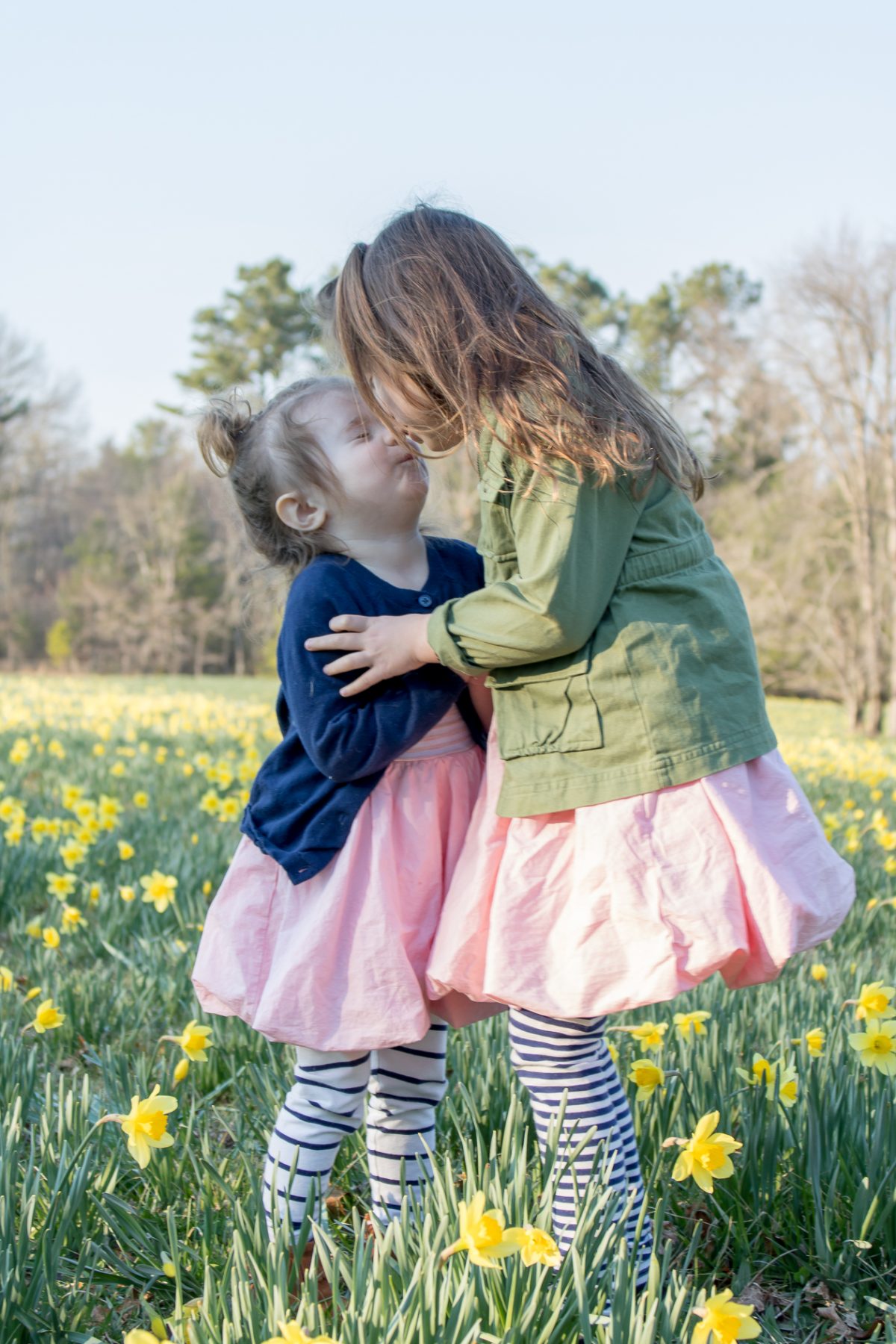 Sisters in a daffodil field, big sister wearing a green coat and pink dress kissing little sister on the nose 