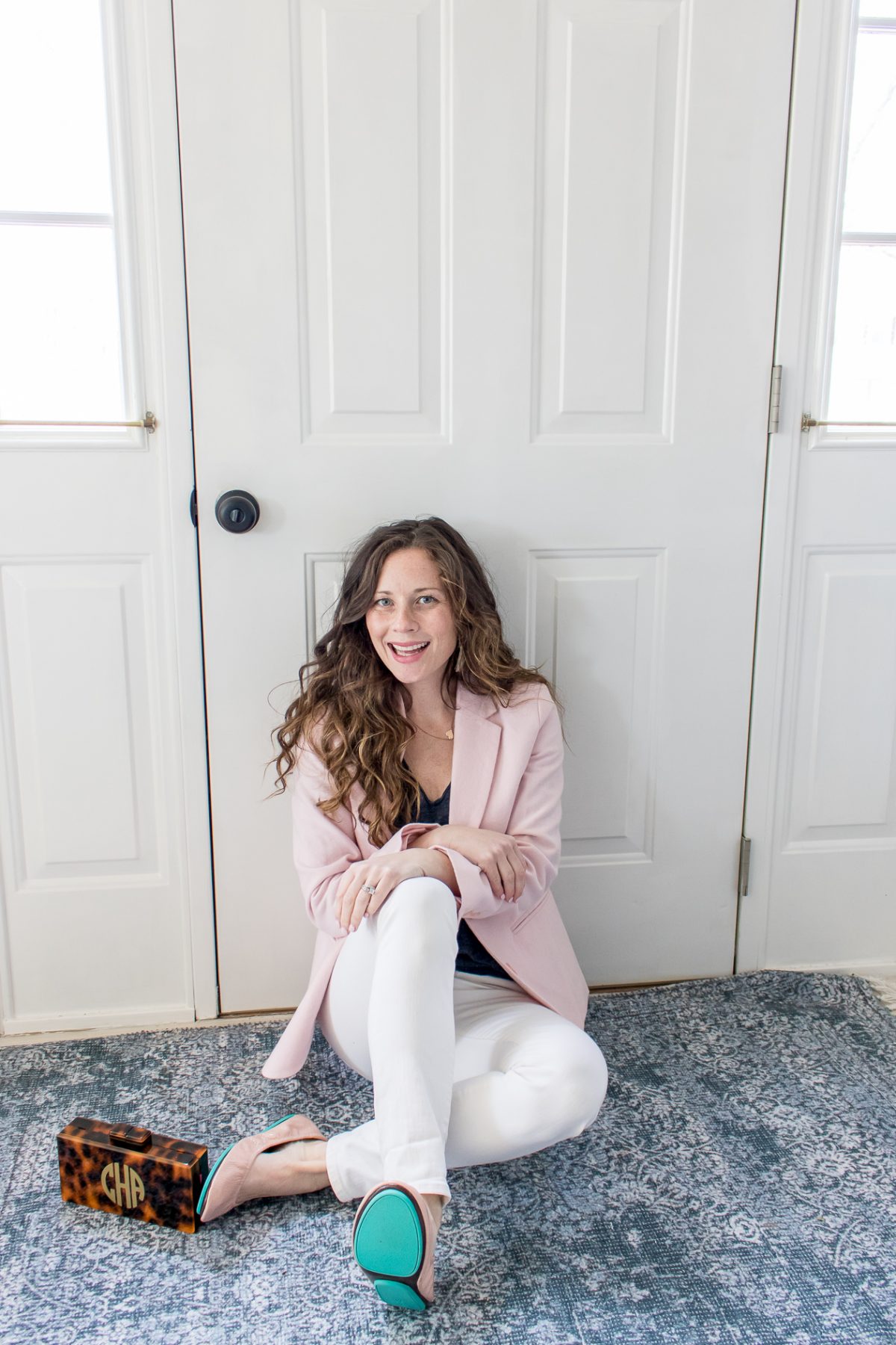 Smiling Woman in pink blazer and white pants sitting in front of a white door on a blue rug with the heel of her blue Tiek Ballet Flats showing