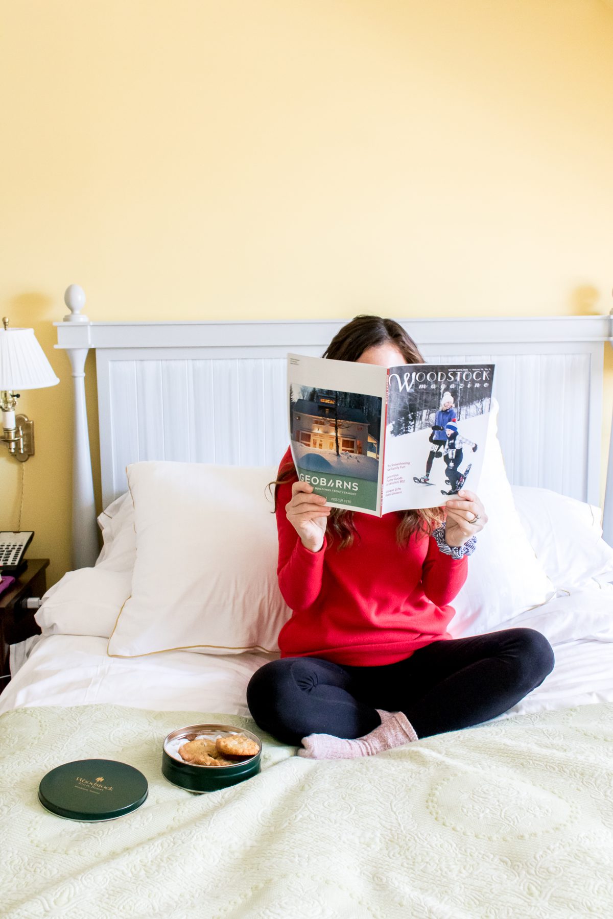 Woman reading Woodstock Vermont magazine on a bed with a tin of cookies in a yellow room