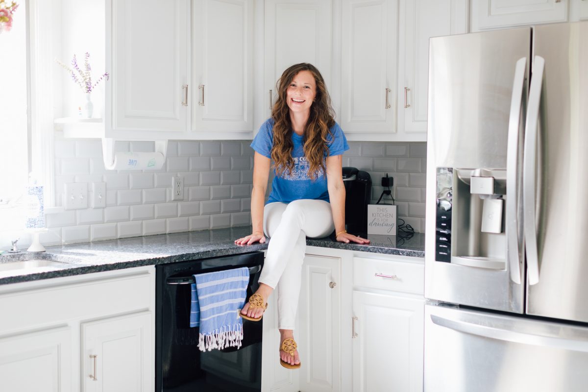 Smiling woman in blue shirt sitting on black countertop in white kitchen