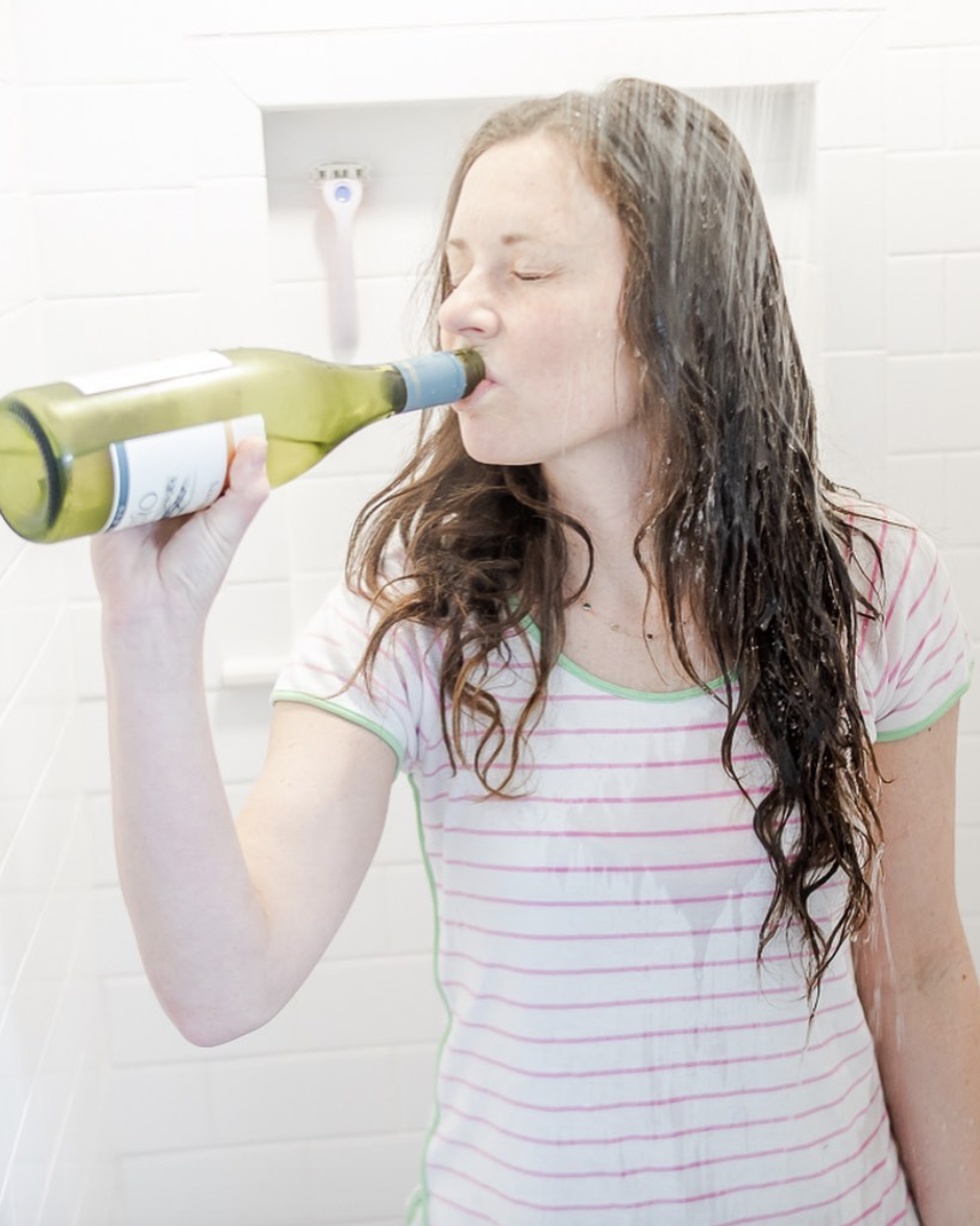 Woman drinking white wine in running shower wearing wearing pajamas 