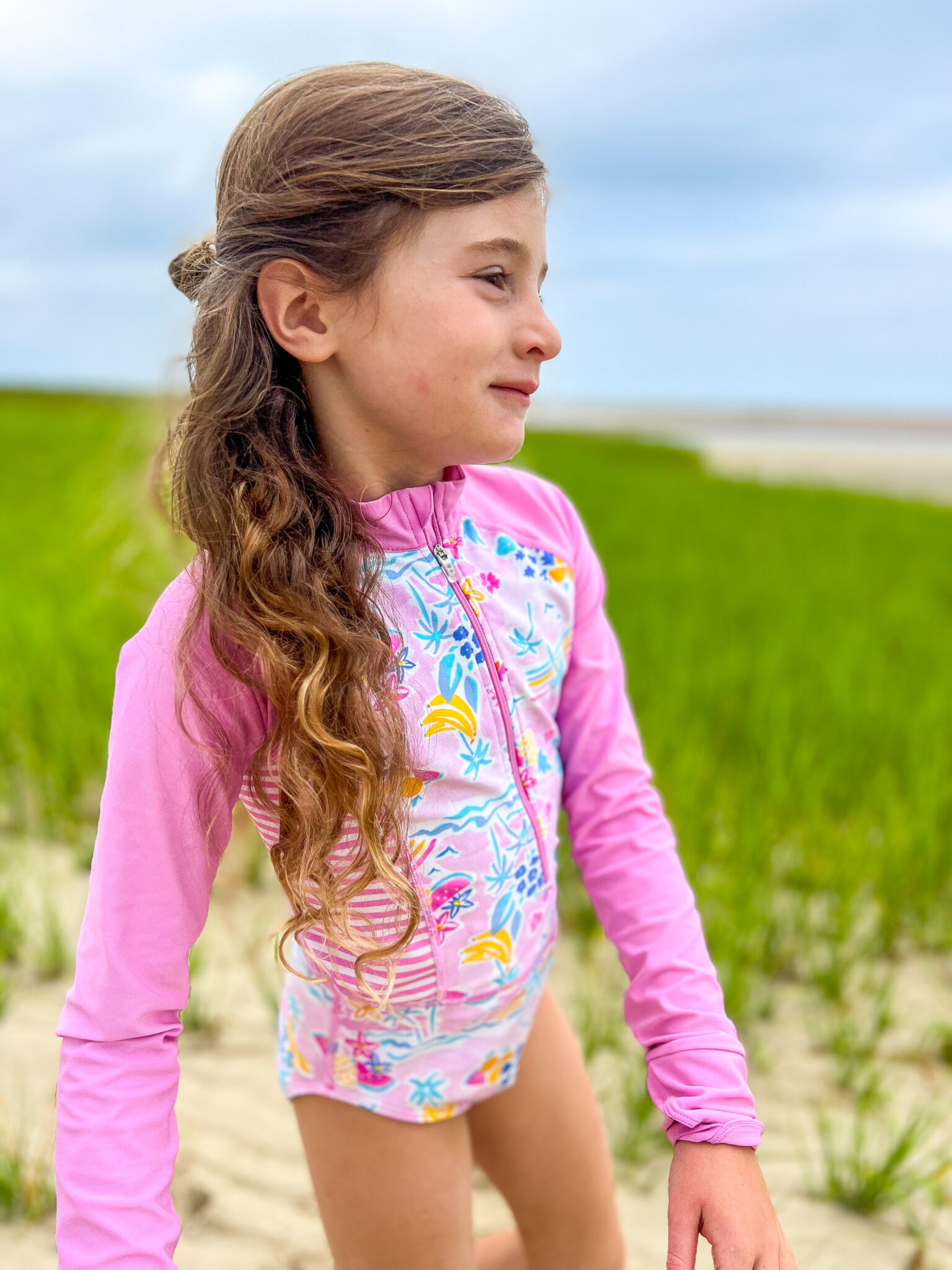 7 year old girl pink bathing suit standing on beach sea grass behind her 