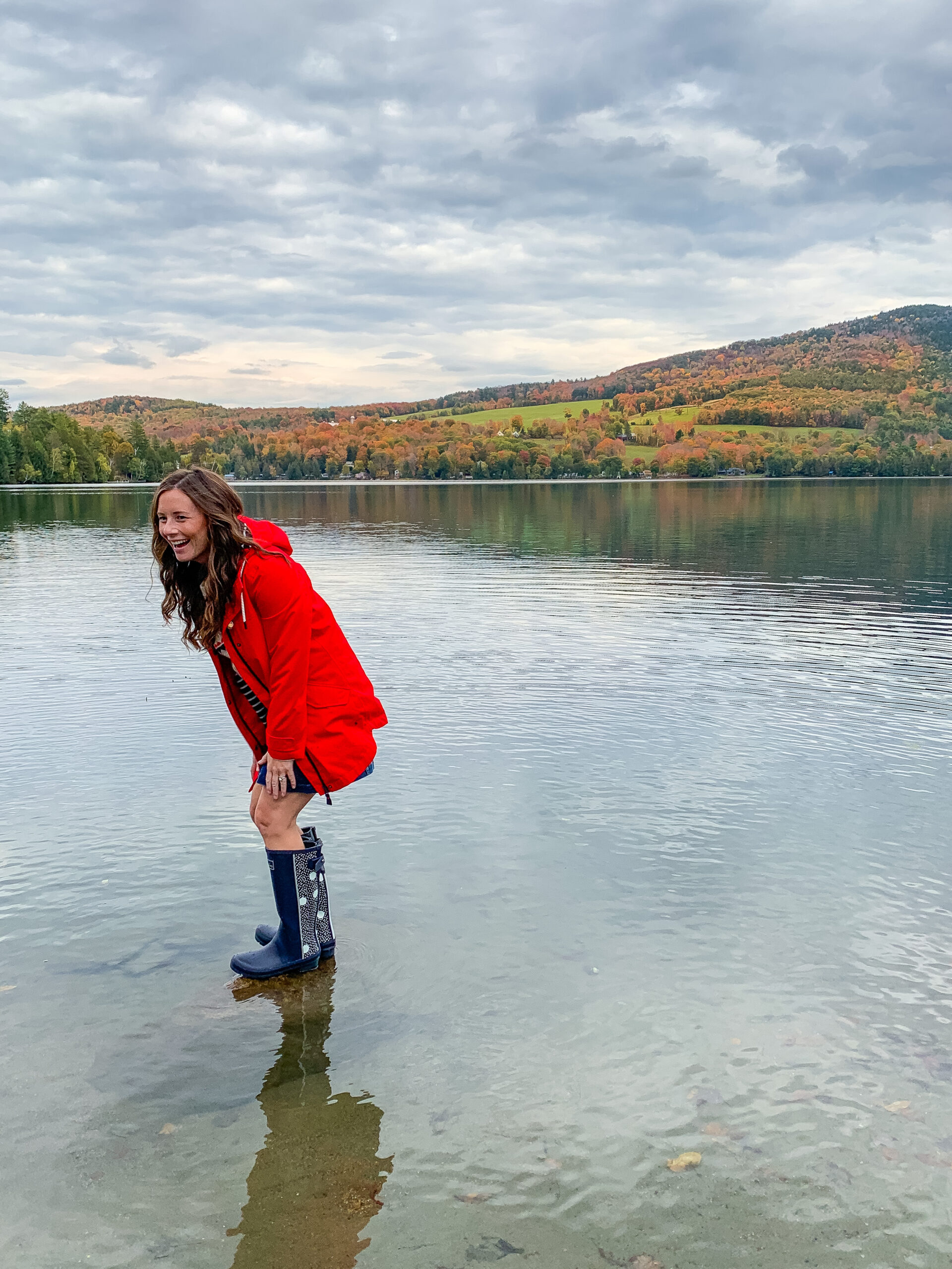woman in rain boots red coat standing in lake in front of fall foliage connecticut 