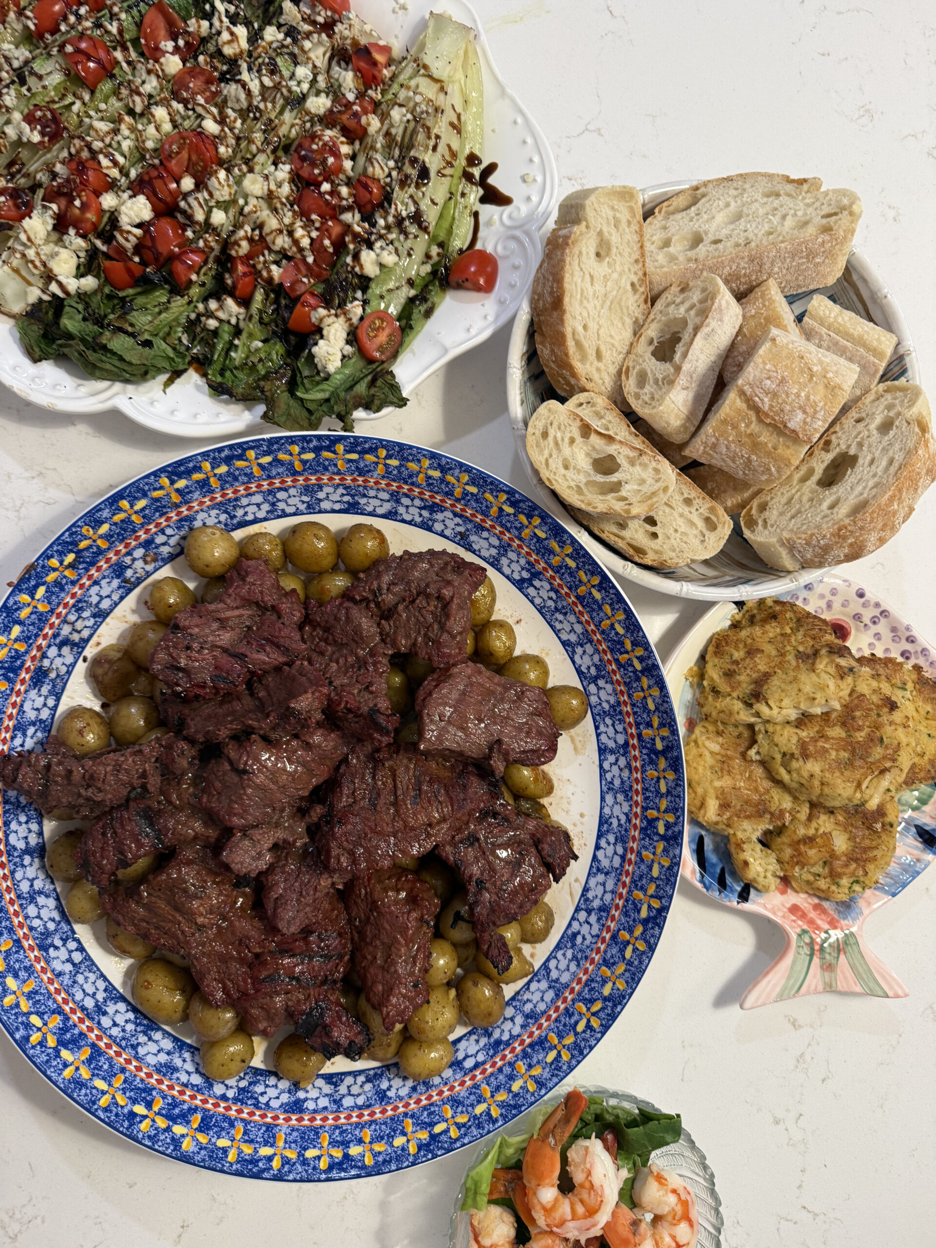 A dinner spread featuring grilled romaine salad topped with gorgonzola, tomatoes, and balsamic glaze; marinated steak tips with green olives on a blue patterned platter; sliced crusty bread; crab cakes; and a small bowl of shrimp cocktail on a white countertop.