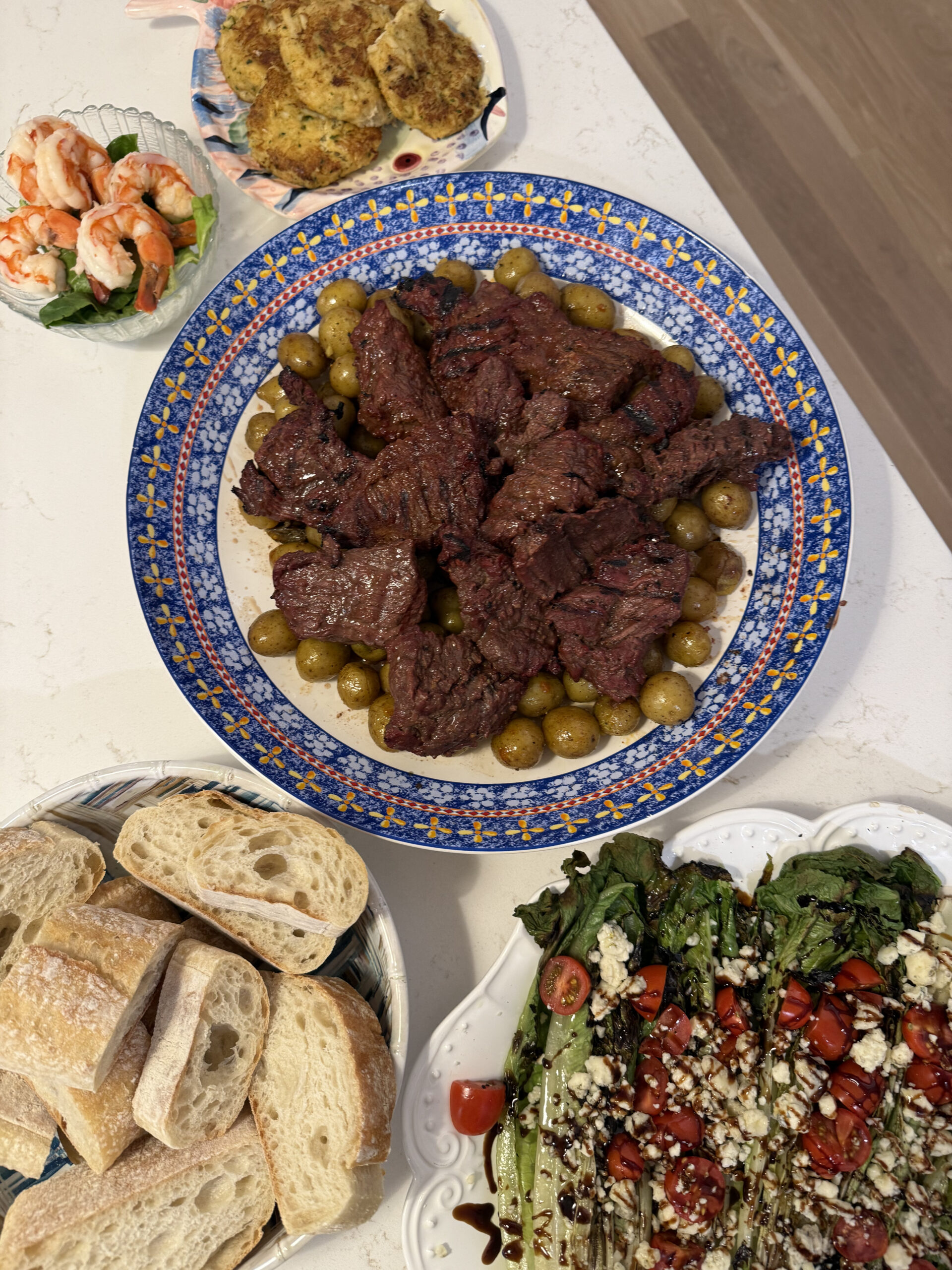 A dinner spread featuring grilled romaine salad topped with gorgonzola, tomatoes, and balsamic glaze; marinated steak tips with green olives on a blue patterned platter; sliced crusty bread; crab cakes; and a small bowl of shrimp cocktail on a white countertop.