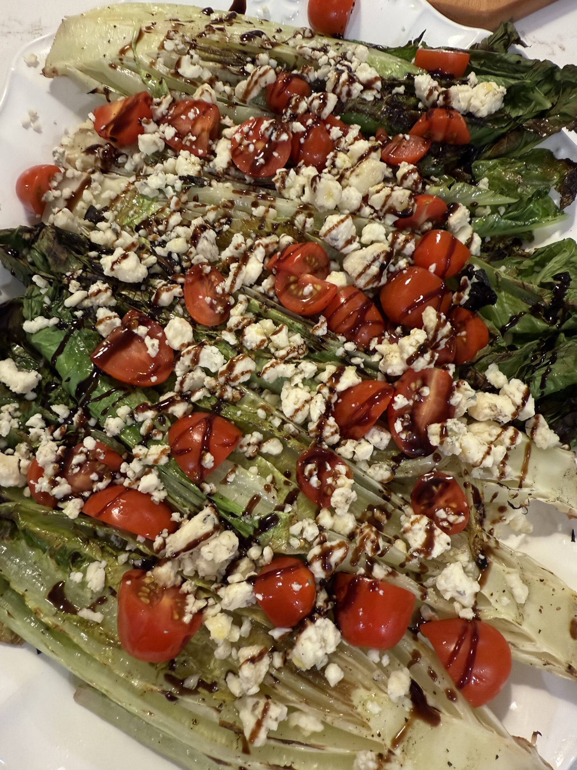 A dinner spread featuring grilled romaine salad topped with gorgonzola, tomatoes, and balsamic glaze; marinated steak tips with green olives on a blue patterned platter; sliced crusty bread; crab cakes; and a small bowl of shrimp cocktail on a white countertop.