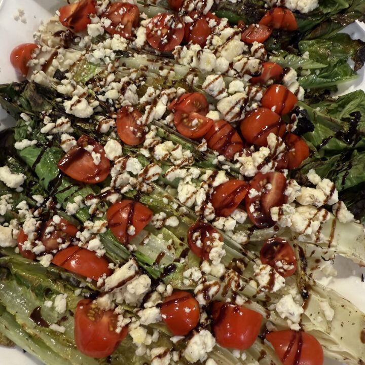 Grilled romaine salad topped with gorgonzola, cherry tomatoes, and balsamic glaze on a white platter.
