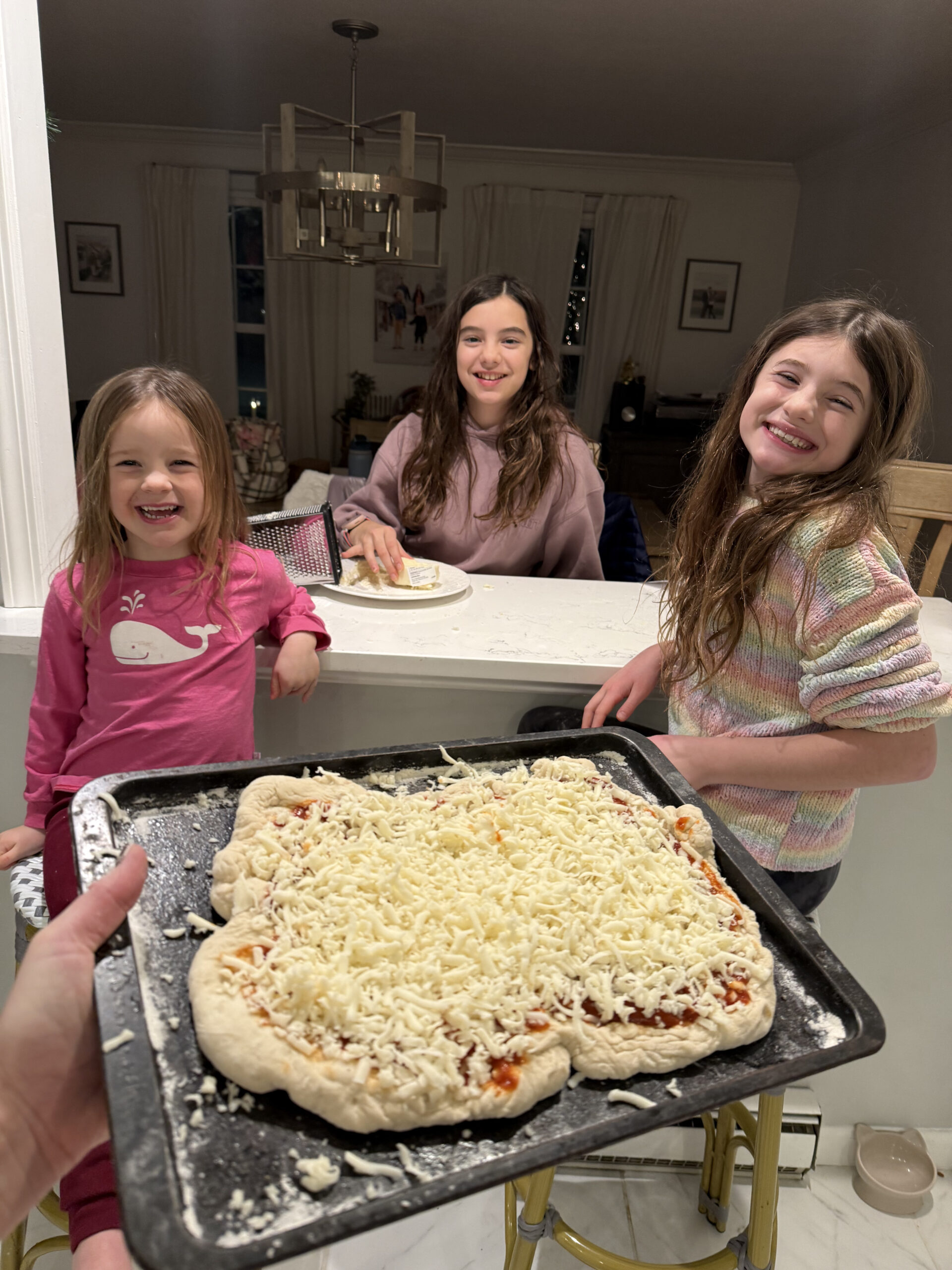 Three girls smiling at camera while Mom holds homemade cheese pizza out to them