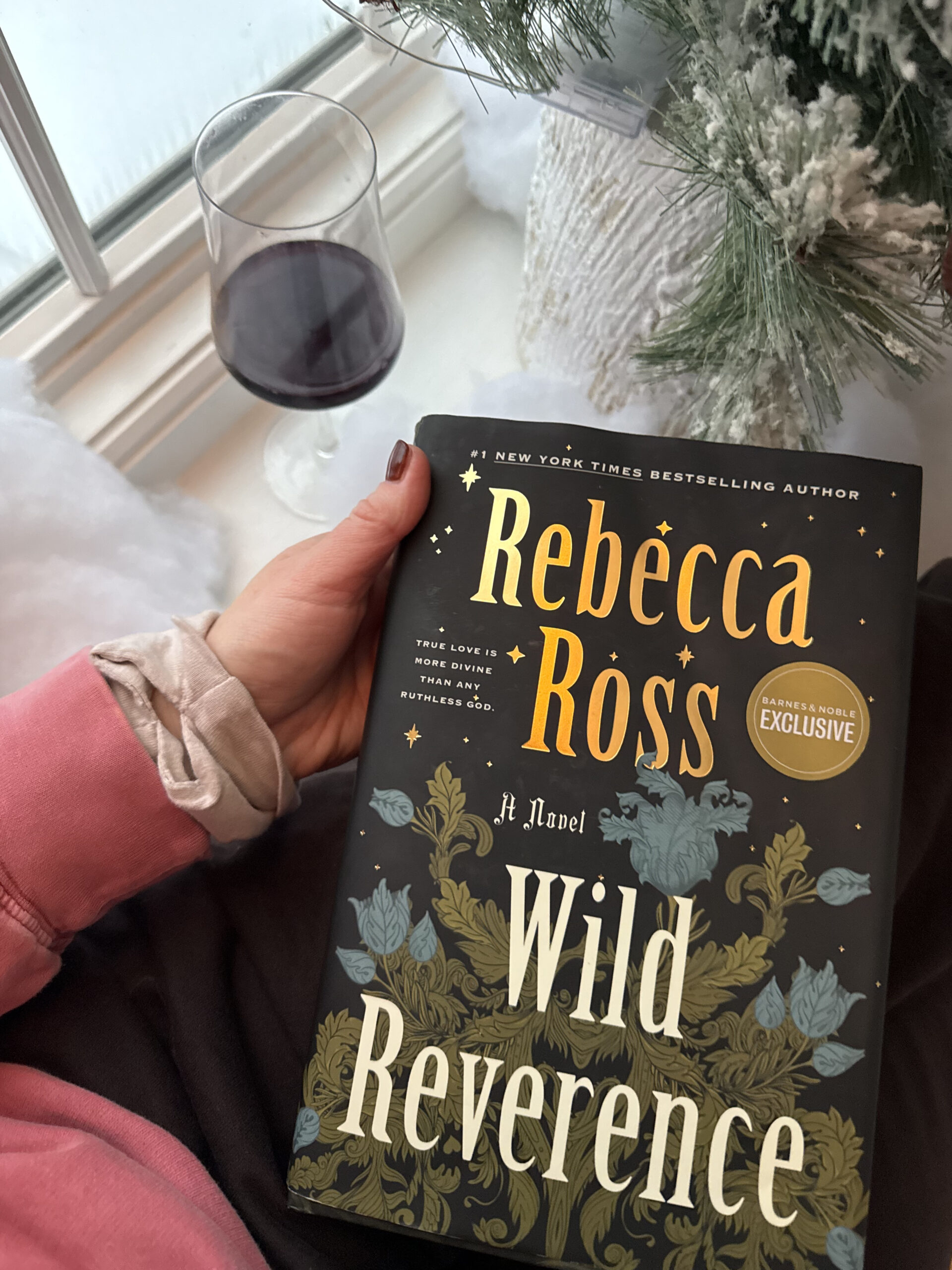 woman holding the book by Rebecca Ross Wild Reverence by a window with a glass of wine